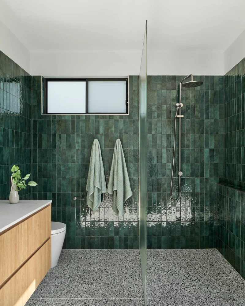 Brass shower fixtures against green tile beside a timber vanity